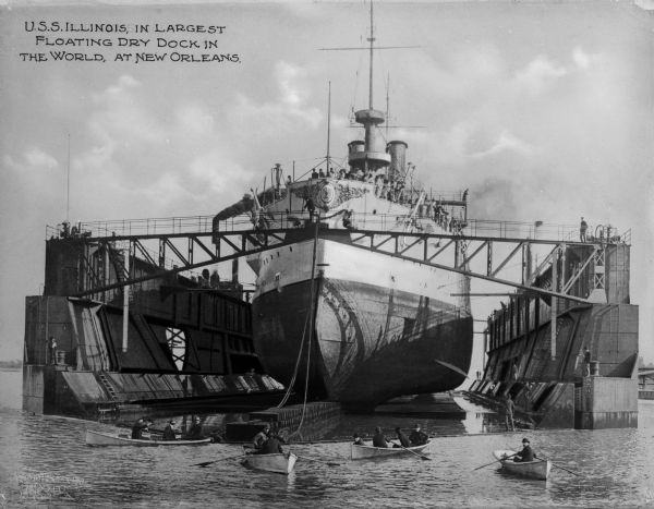 The USS <i>Illinois</i> in the largest floating dry dock in the world at the time, surrounded by rowboats in the foreground. The battleship was built at Newport News, Virginia, and was one of three 11,565-ton battleships. She was commissioned in September 1901. One of her first assignments was to test the newly-constructed floating dry dock at the Naval Station. Text on photograph reads: "U.S.S. Illinois, in largest floating dry dock in the world, at New Orleans."