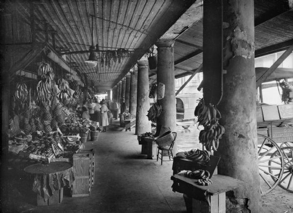 View of colonnade and French market in New Orleans, Louisiana. Pineapples and bananas are hanging in the stalls and in the foreground near a column.