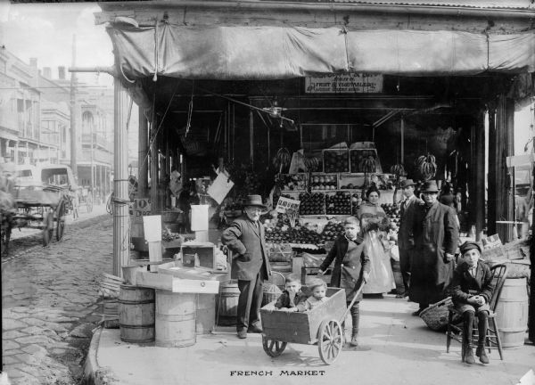 View of shoppers in the French market. One boy is shown pushing two small children in a cart. Fruits and vegetables can be seen in the market stall in the background. Caption reads: "French Market."