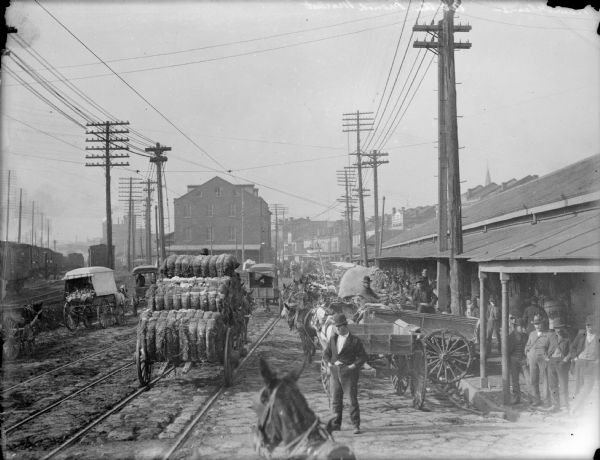Slightly elevated view of the French market. A cart transporting bales of cotton is in the street on the left. Railroad tracks are in the street.
