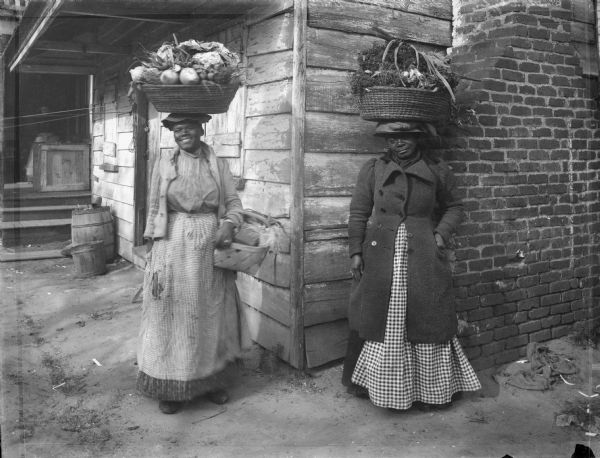 Two African American women pose with baskets of fresh vegetables on their heads while selling their produce. A building with an exterior brick chimney is behind them.