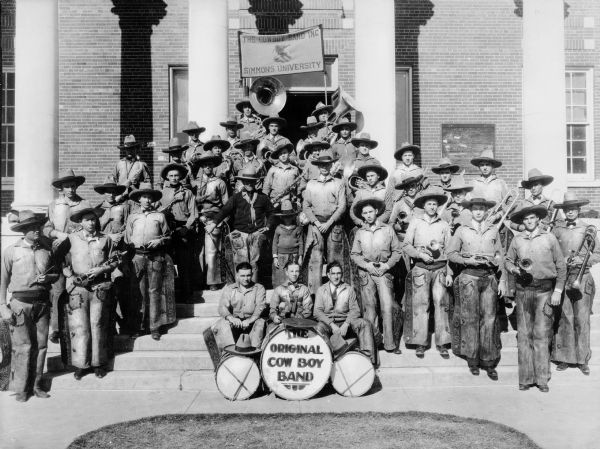 The Original Cowboy Band poses on the steps of Simmons University in Abeline, Texas. Writing on drum reads: "The Original Cow Boy Band" and writing on banner reads: "The Cowboy Band Inc. Simmons University."