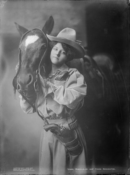 A cowgirl, outfitted with her holstered pistol and standing by a horse. Caption reads: "The Belle of the Ranch."