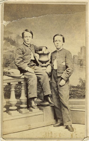 Carte-de-visite full-length studio portrait of Charles David and Harrie Farwell Atwood, the sons of David Atwood of Madison, Wisconsin. They are posed on and next to a prop balustrade in front of a painted backdrop.