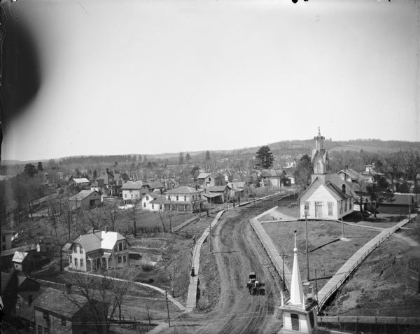 Elevated view of the street and board sidewalk. Robie house located lower left; Charles Van Schaick residence with porch located in the center; Roman Catholic Church to the right. In foreground is the Baptist Church, later St. John's.