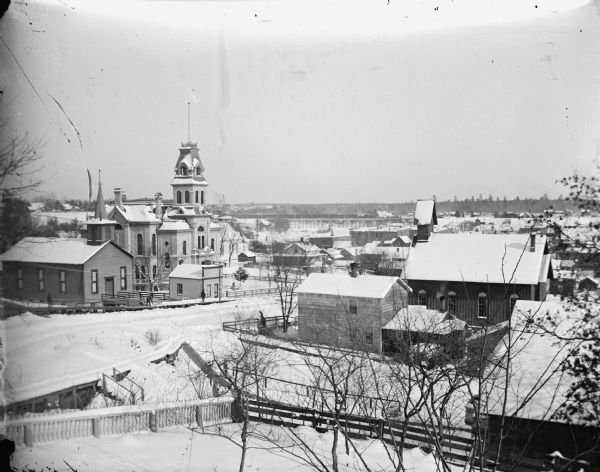Elevated view, with snow on the ground, of Main Street with the Baptist Church on the left facing the Episcopalian Church at the right center. Center left is the Jackson County Courthouse, which obscures the Norwegian Lutheran Church. Foreground, home of Lew Dimmick, a harness-maker and Civil War Veteran who lost his leg. Further up the hill, Main Street leads to the Universalist Church, the Catholic Church, and the home of Charles J. Van Schaick.
