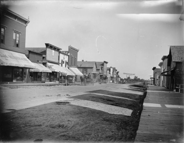 South side of Main Street from the intersection with Merrill Street. Various storefronts are on the left, including J.M. McCormick Dry Goods and Groceries; Syd. E. Brown Millinery; City Cash Grocery; a restaurant; and the Exchange Hotel; the City Bakery on the other side of Main Street.