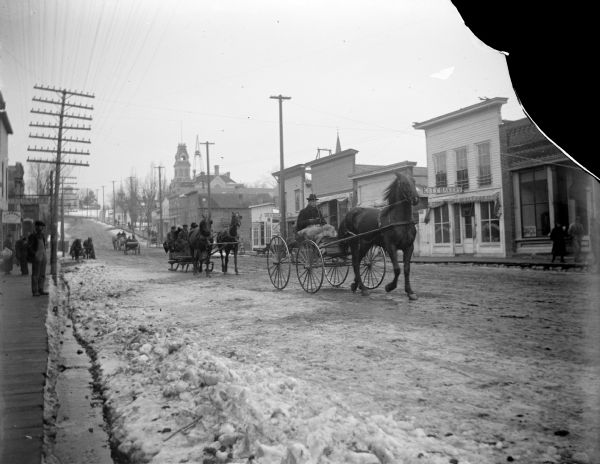 Wagons and sleighs on a snow-covered Main Street. Looking west on Main Street toward the intersection of the Main Street and First Street. Storefronts, from left to right, include a Hardware store and the City Bakery.