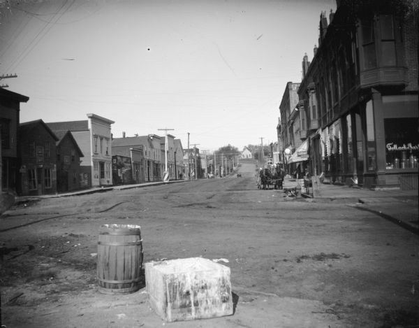 Barrel and box on the Main Street, looking west up the street. Large barber pole on the left side up the street, and Tollack and [?] storefront on right street corner.
