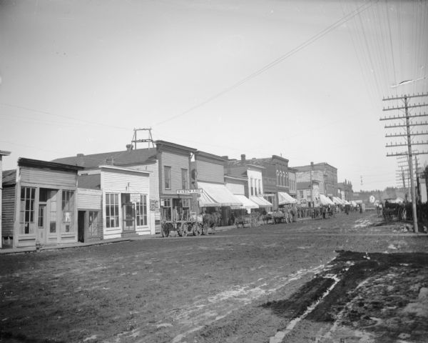 Main Street lined with horses and wagons, with a large crowd of people gathered at the intersection. Storefronts, from left to right: City Barbershop, owned by H.A. Johnson; a Hardware Store; G.G. Melrose (?) Crockery; Sprester Bros. Groceries, and City Bakery.