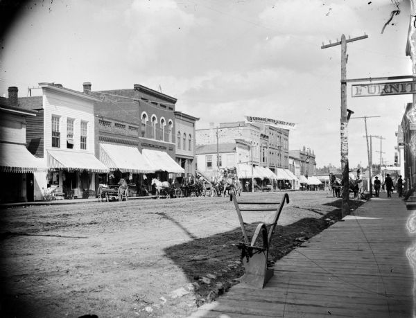Main Street storefronts, identified from left to right as: The Fair ["Bargains in Everything" on the awnings], and Eckern Jeweler. Banner across Main Street advertises the "La Crosse Inter State Fair, Sept. 14, 15, 16, and 17, Admission 25 Cents". In the foreground a plow sits on the board sidewalk, near a furniture store. Horses and carriages are in the street.