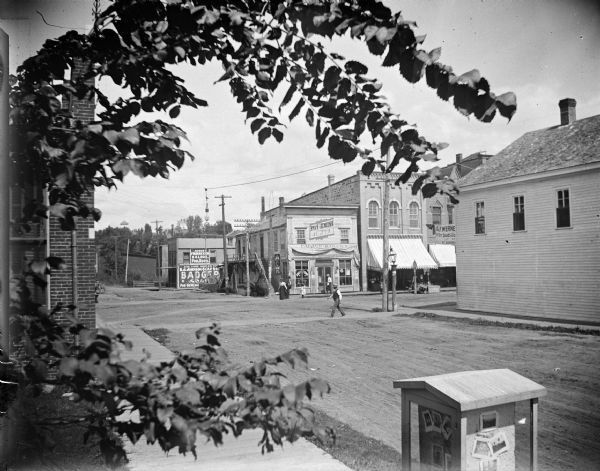 Intersection of Main and First Streets, showing storefronts including, from left to right, August Frederickson Saloon/Poolroom, Palace Bakery/American Express Company, A.F. Werner. Lower right foreground is a display case of Van Schaick photographs. Banner across Main Street reads "Bridge Park, Aug. 12".