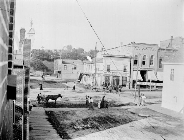 Elevated view of busy intersection of town, looking northeast across Main Street on First Street. Persons walking, and horses and carriages. Storefronts include, from left to right, the Palace Bakery and Confectionery, with a window for A.L. Werner; A.F. Werner Store; and a sign on the building on the southwest corner for "Milwaukee Beer".