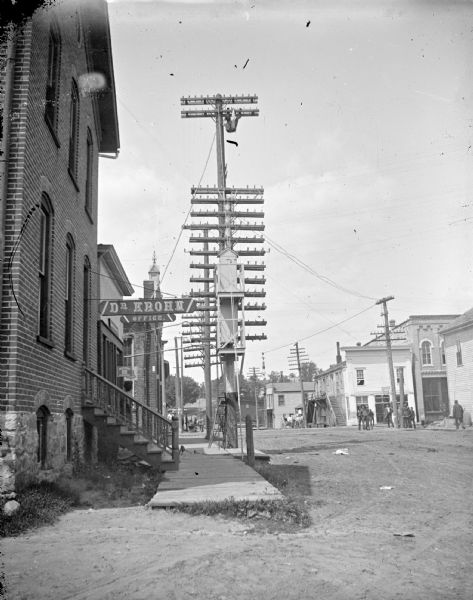 South First Street and the intersection with Main Street. Men on corner looking at man on telephone pole. The man hanging from the pole is probably Ernest Manthe. Signs on the buildings on the right side of First Street are for Dr. Krohn's Office. The building on the corner with the alley belonged to Charles J. Van Schaick and housed the telephone exchange and the office of Dr. Eugene Krohn.