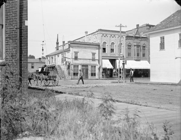 Intersection of Main and First Streets, shows southwest section of intersection, with street lamp, storefronts, pedestrians, and carriage.  Taken from Empty Lot on South First Street, between the Studio and Bank. Storefronts visible include, from left to right, building with painted windows for "A.F. Werner" and "Gents Furnishings"; unidentified store, and Smolensky New Dry Goods.