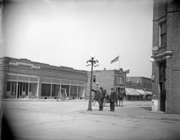 Man leaning on electric street light. Buildings and ornamental street light show post-flood developments. Building on northwest corner of intersection in process of construction. The lamp post is on the southeast corner of the intersection of Main and South First Streets. First storefront awning displayed in the right side of image is for L.H. Hagen and Son.