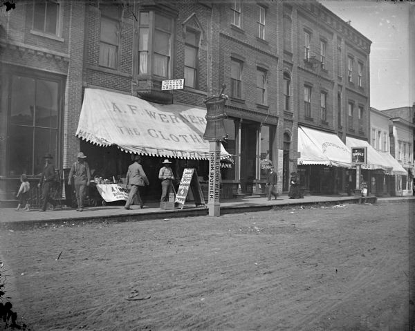 Families gathered outside of Werner's Drugstore on annuity pay day. A sign reads "Medicine sold here" in Ho-Chunk, German, and Norwegian. The Ho-Chunk syllabary on the sign "Ma Ko Oro Wena" reportedly translates colloquially as "medicine/place where you buy" and Norwegian "Norsk Apothek."