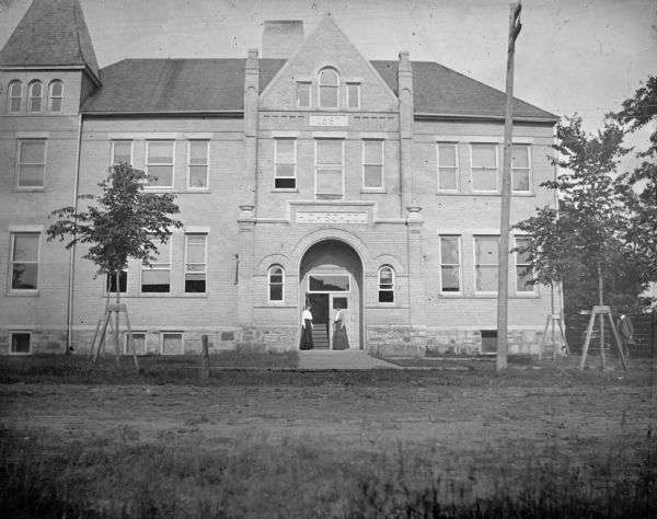 View across yard towards the old High School Building, with two unidentified women posing standing in the doorway arch, and an unidentified man standing on the far right.