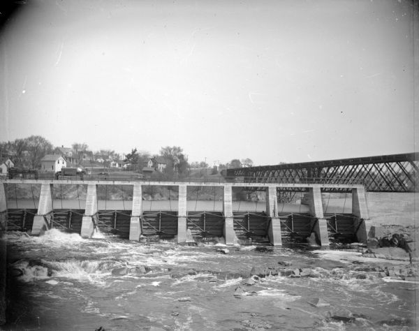 One side of a concrete dam with a railroad bridge to the right, and houses in the background.