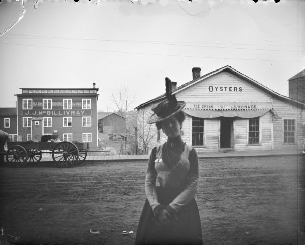 Woman in a tilted hat posing in the foreground. Across the street are stores: J.J. McGillivray Sash and Door Company and Squire's Oyster House. Probably on Water Street.