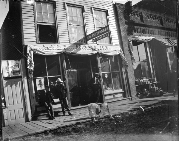 Three men leaning and standing in front of the City Bakery. Large blocks of ice on the board sidewalk. Poster in the window is advertising the Jackson County Fair on September 19, 20, 21, 22.