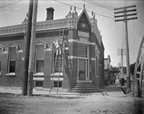 Man on a ladder painting around a window of the Jackson County Bank. Arch over the intersection partially visible on the right side, as are several people on the south sidewalks of Main Street. There are signs for "Furniture" and "Undertaking".