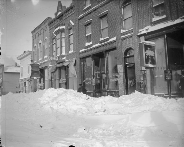 Winter scene with Main Street, north side, between First and Water Streets, Black River Falls, Wisconsin, piled high with snow. Mortar and pestle sign is in front of the Werner's Drugstore. The next building has a sign for Dr. Abbott, with a man standing in front of the First National Bank. The store on the far right has a sign for "The People's Drugstore" and "Apothek". The shadow of the photographer is on the snow bank in the lower right corner of the image.