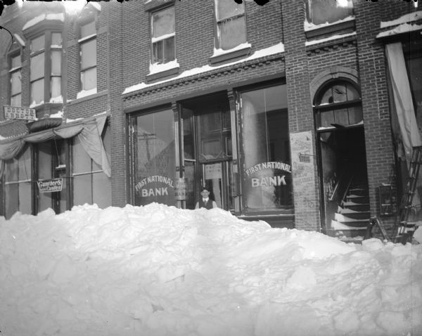Winter scene of a snowbank in the middle of the north side of Main Street between Water and First Streets, Black River Falls, Wisconsin. Storefronts identified from left to right include Dr. Abbott and the First National Bank.