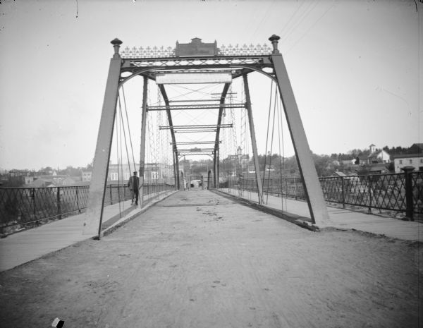 Bridge with metal framework and sidewalks on each side. Man walking away from the camera on the left sidewalk. Sign across the bridge, "$10. FINE FOR DRIVING [ACROSS] THIS BRIDGE FASTER THAN A WALK." Bridge constructed according to the upper plate by "[?] & Iron Works."