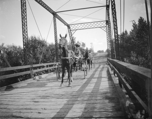 Horses and wagons passing over a plank bridge. Woman driving the lead wagon pulled by a single horse, and a man and another obscured person in the second wagon pulled by two horses.