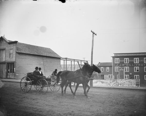 Two wagons and teams driving past the J.J. McGillivray Sash and Door Company. Two men in the wagon pulled by two horses in the foreground, and a man in the wagon pulled by a single obscured horse. Billboards advertise the "Georgia Up-To-Date Minstrels" at the Opera House on April 7.