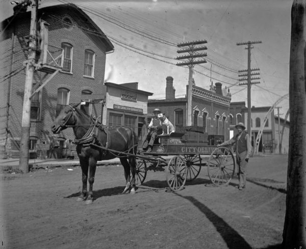 Boy driving the City Delivery wagon pulled by a single horse and accompanied by a white dog. A man is standing next to the wagon. Storefronts include, from left to right: the Van Schaick Building, with the American Express Office and Dr. Penton; J.R. McDonald, and the Jackson County Bank on the corner with Main Street.