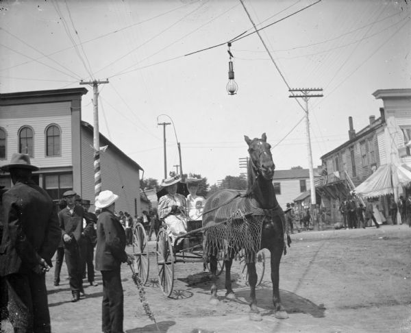 Two women, one holding a child, in a wagon pulled by a single horse wearing flynets. There are several people in the intersection, during what looks to be a patriotic celebration.
