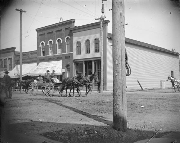 Native American man, and two women in a wagon pulled by a single horse. Storefronts include a jeweler on the left with an awning advertising books and stationery, and a small sign for J.R. [?] Perry, Lawyer.