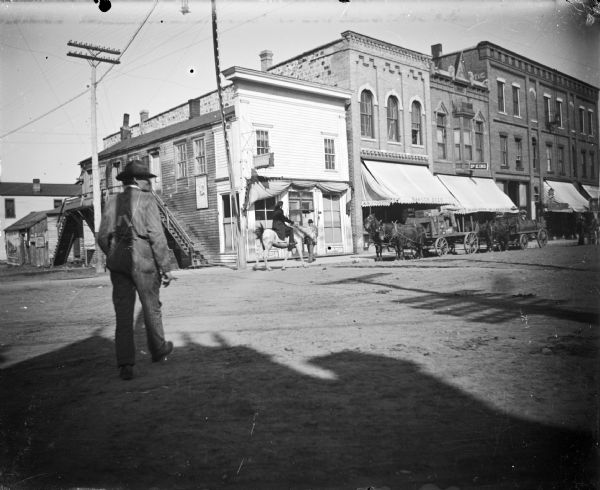 Buildings on the southeast section of the intersection of Main and First Streets. Two freight wagons with teams of two horses in front of stores. Man crossing street away from camera, and a Ho Chunk man on a light-colored pony in the background. Storefronts identified from left to right as, a saloon, store of Moses B. Paquette and awning advertising Bangor Beer, and the A.F. Werner store with awning advertising "Bargains in Everything" below a sign for Dr. King.