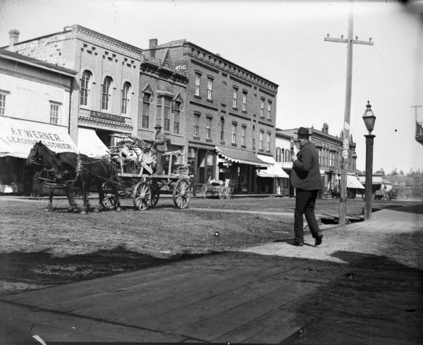 Wagon piled high with chairs, with a man standing and driving a team of two horses. A man on the sidewalk is looking back at camera. There are other wagons in the street in the distance on the right side. Storefronts identified, from left to right, are the store of A.F. Werner, A. Meinhold, obscured, First National Bank, and the rest unidentified.