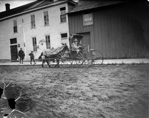 A "cart before the horse" rig advertising the "A.F. Werner, Bargains in Everything, Black River Falls, Wis.", with a man wearing a fake bushy beard and holding a trumpet, and children on the sidewalk next to the rig. A man wearing a military uniform is walking on the sidewalk behind the cart and carrying a French horn.