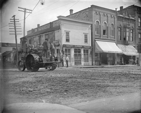 Two men driving a steam tractor in town with a few spectators. Storefronts, identified from left to right, including, O.J. Heath Saloon, store of Moses Paquette with an awning advertising Bangor Beer, and A.F. Werner under a sign for Dr. Abbott.