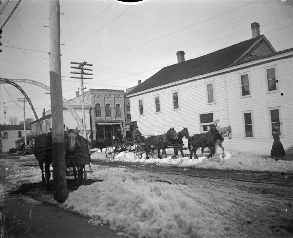 Winter scene looking toward Main Street from South First Street, with an arch over the intersection. A man is operating a road grader pulled by four horses across the street plowing snow, and a wagon pulled by two horses is tied to a pole on the left. There is a woman walking on the sidewalk on the far right. Awning advertising Bangor Beer at Moses Paquette is on Main Street.