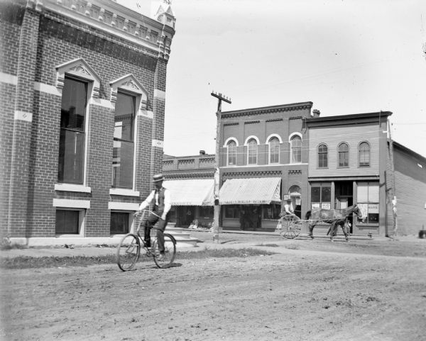 Man riding bicycle down First Street near Main Street. In the background is a man driving a horse and two-wheeled buggy on Main Street. Storefronts identified on Main Street include, from left to right, Eckern Grocer (partially visible), Chicago Cheap Store, a lawyer's office, Journal Office, and a hardware store (probably named Murphy).