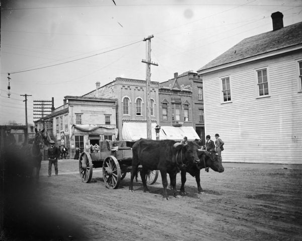 Oxen in yoke pulling an empty wagon at intersection of First and Main Street. Storefronts identified on the northeast corner of Main and First Streets include, from left to right: Palace Bakery and American Express Office, A.F. Werner, ? Store with "Bargains in Everything" and Dr. Abbott, and the First National Bank.