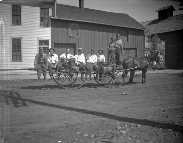 View across unpaved street towards six men posing sitting, and a boy and a man posing standing on a wagon pulled by a single horse. One man is obscured standing behind the horse. Tobacco advertisements for "Tom Keene Cigars", "Bull Durham Tobacco", and "George 5 cent Cigars" on the small building on the far right.