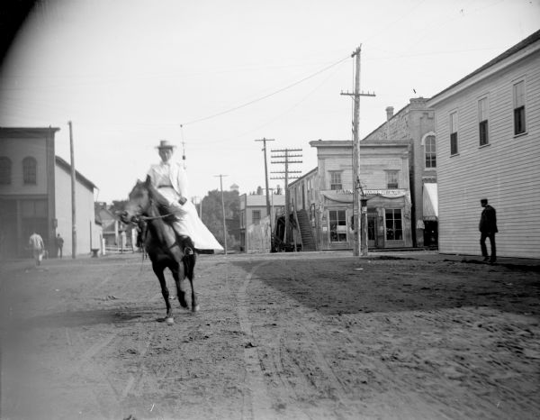 Woman riding sidesaddle through town. Storefront on the northeast corner of Main and First Streets as the Palace Bakery and American Express Office.
