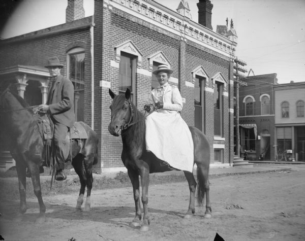 Outdoor view towards a woman and man posing on horseback. The woman is riding sidesaddle. Storefronts identified on Main Street, from left to right, as J.A. Eckern Jeweler and P.L. Moe & Company Hardware.