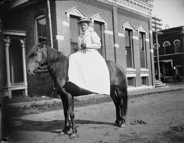 View of a woman posing sidesaddle on a horse. Storefront on Main Street identified as J.A. Eckern Jeweler.