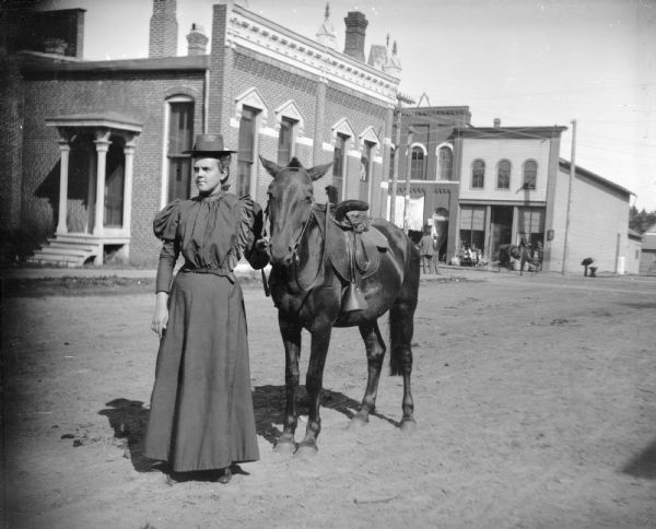 Woman standing and holding saddle horse by reins on street in front of Jackson County Bank on First Street, possibly Edna Bright. P.L. Moe and Co. Hardware Store visible on corner, and the jewelry store is partially visible on its left.