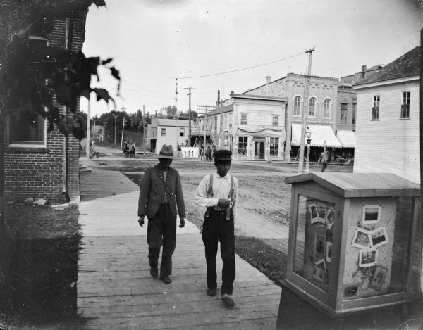 Two boys walking on board sidewalk of First Street away from Main Street. In the right foreground is a display case of Van Schaick photographs, in front of his studio. Identified on the north corner of the intersection, from left to right, is the Bakery and the A.F. Werner Store.