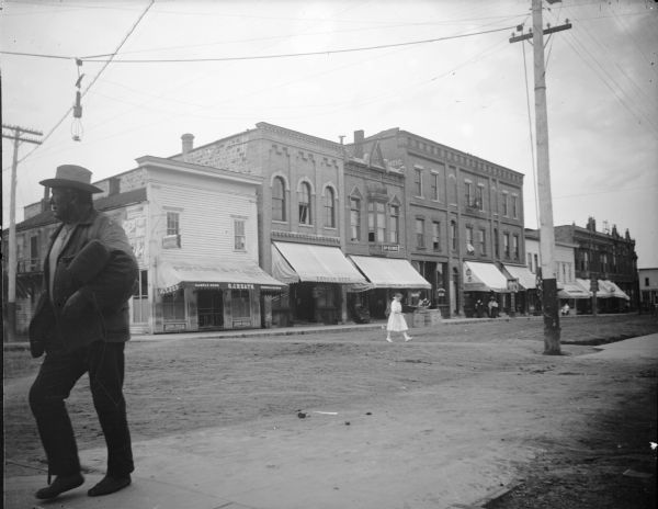 Girl in a light-colored dress walking across Main Street and a man crossing in left foreground. Storefront on Main Street, identified from left to right, O.J. Heath Saloon, Mose Paquette, Werner Drugstore with a sign for Dr. King, the First National Bank, and several unidentified storefronts.