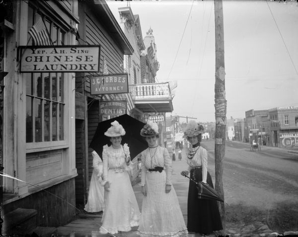 Three women (Jennie Parsons Kelly, left, and Lena Eisenback Post, center, and an unidentified woman on the right) on Main Street in front of Yep Ah Sing's Chinese laundry. They wear elaborate hats and carry parasols. Other signs on the street include J.C. Forbes, Attorney; Dr. J.W. Boisol, Dentist; and City Library.