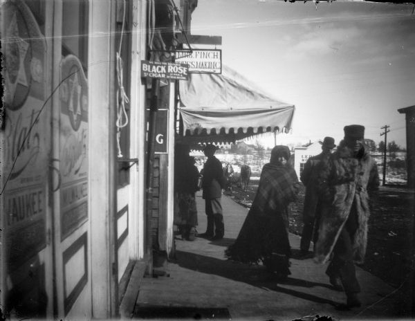 Man wearing a fur coat, a woman wrapped in a blanket, and other individuals bundled for warmth on a winter day. In front of the millenary shop of Miss Finch and a saloon serving Blatz beer. Snow-covered riverbank in background.